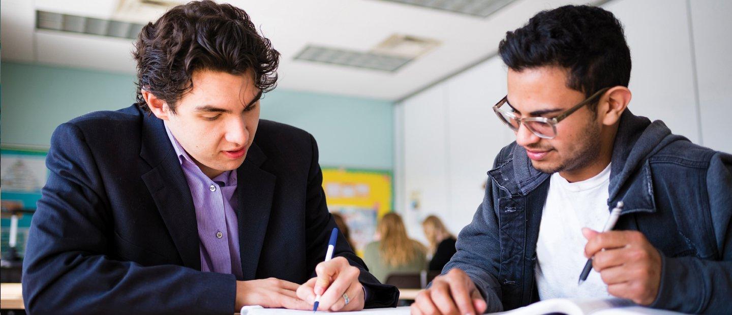 两个年轻人坐在教室的书桌前，在笔记本上写着什么